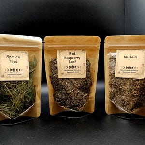 Może przedstawiać: Trzy brązowe torebki z papieru kraft z suszonymi ziołami. Etykiety zawierają napisy Spruce Tips, Red Raspberry Leaf oraz Mullein, grafikę faz księżyca oraz tekst Moon Phase Naturals Harvested Herbs.