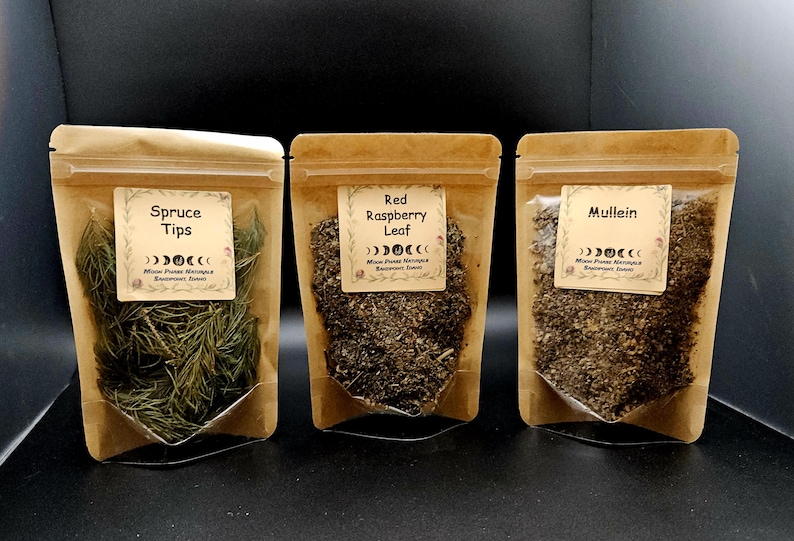 Może przedstawiać: Trzy brązowe torebki z papieru kraft z suszonymi ziołami. Etykiety zawierają napisy Spruce Tips, Red Raspberry Leaf oraz Mullein, grafikę faz księżyca oraz tekst Moon Phase Naturals Harvested Herbs.