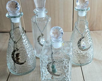 May include: Four clear glass bottles with faceted crystal-style stoppers. Each bottle features a silver-toned chain necklace draped around the neck, adorned with a crescent moon charm and small hanging beads or stones in various colors.
