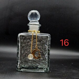 May include: A clear glass perfume bottle with a square body featuring a textured, pebbled surface. The bottle has a faceted, diamond-shaped stopper and a gold-toned chain charm with a crescent moon, a small Eiffel Tower, and a white bead. The number 16 is visible.