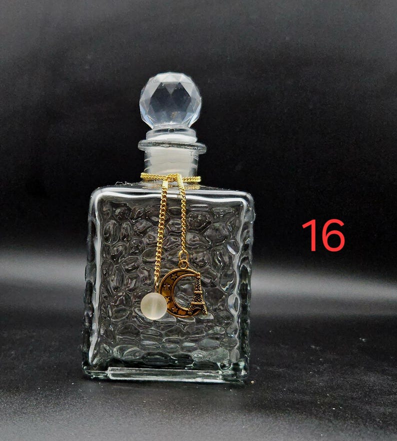 May include: A clear glass perfume bottle with a square body featuring a textured, pebbled surface. The bottle has a faceted, diamond-shaped stopper and a gold-toned chain charm with a crescent moon, a small Eiffel Tower, and a white bead. The number 16 is visible.
