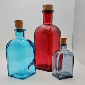 May include: Three coloured glass bottles with cork stoppers. The bottles are blue, red, and grey, and vary in size and shape. The bottles have a square base and rounded shoulders.