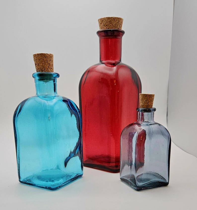 May include: Three coloured glass bottles with cork stoppers. The bottles are blue, red, and grey, and vary in size and shape. The bottles have a square base and rounded shoulders.