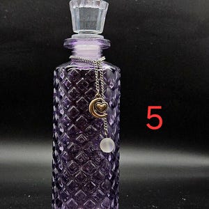 May include: A purple glass bottle with a diamond-patterned texture and a clear glass stopper. A silver-toned chain hangs from the neck, featuring a gold-colored crescent moon charm with a small heart and a translucent white spherical bead. The number 5.