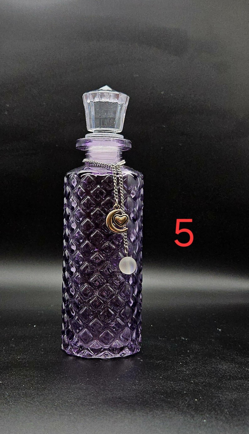 May include: A purple glass bottle with a diamond-patterned texture and a clear glass stopper. A silver-toned chain hangs from the neck, featuring a gold-colored crescent moon charm with a small heart and a translucent white spherical bead. The number 5.
