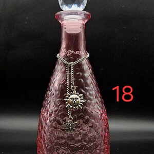 May include: A pink glass bottle with a textured, dimpled surface and a clear, faceted crystal-style stopper. A silver-toned chain hangs around the neck, featuring a sun face charm and a small butterfly charm. The number 18 is visible in red.