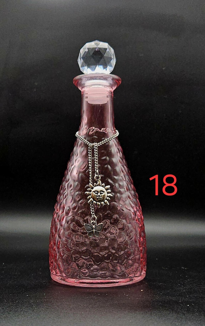 May include: A pink glass bottle with a textured, dimpled surface and a clear, faceted crystal-style stopper. A silver-toned chain hangs around the neck, featuring a sun face charm and a small butterfly charm. The number 18 is visible in red.