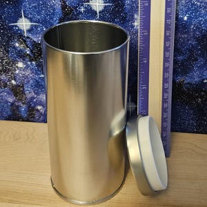 Może przedstawiać: Cylindryczny srebrny metalowy pojemnik z otwartą g&oacute;rą i pasującą pokrywką z białym plastikowym brzegiem. Drewniana linijka umieszczona obok pojemnika wskazuje wysokość około 19 cm dla tego naczynia.