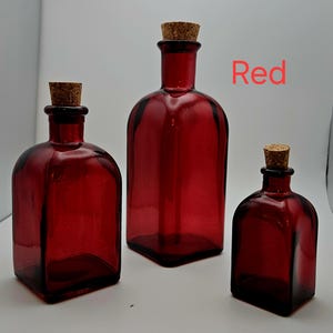 May include: Three red glass bottles with cork stoppers. The bottles are different sizes, with a square base and rounded shoulders. The word "Red" is written in red text.