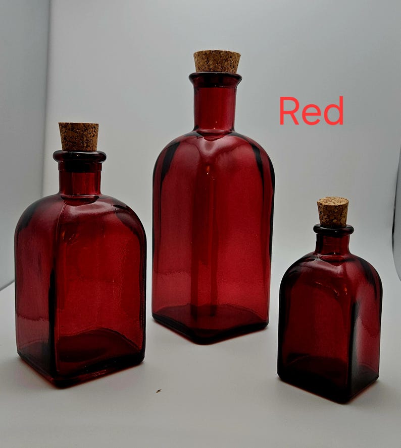 May include: Three red glass bottles with cork stoppers. The bottles are different sizes, with a square base and rounded shoulders. The word "Red" is written in red text.