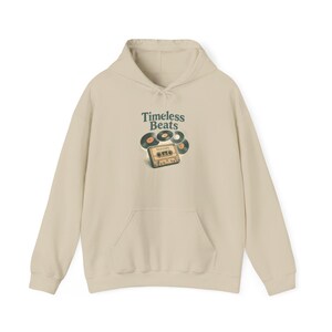 Può includere: Una felpa con cappuccio beige con tasca frontale e cappuccio con coulisse. La parte anteriore della felpa con cappuccio presenta il testo "Timeless Beats" sopra una grafica di dischi in vinile e una musicassetta.