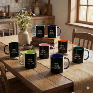 May include: A collection of black ceramic mugs with colorful interiors and handles, including blue, purple, orange, green, and pink. Each mug features the text "PRAY. TRUST. EXECUTE." on the front, set on a wooden table.