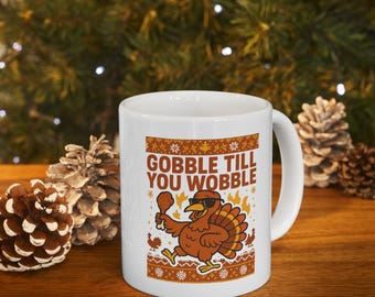 Può includere: Tazza in ceramica bianca a tema Ringraziamento. La tazza presenta un tacchino dei cartoni animati con occhiali da sole e un cosciotto di pollo, con la scritta "GOBBLE TILL YOU WOBBLE". Il design include una combinazione di colori marrone e arancione.