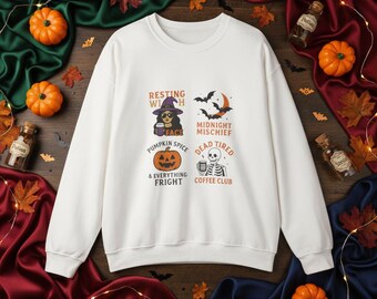 May include: A white sweatshirt with Halloween-themed graphics. The design includes a witch, bats, a jack-o'-lantern, and a skeleton. Text on the shirt reads "Resting Witch Face", "Pumpkin Spice & Everything Fright", "Midnight Mischief", and "Dead Tired Coffee Club".