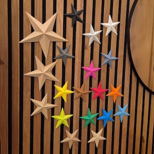 Könnte beinhalten: Eine Sammlung sternförmiger Wanddekorationen in verschiedenen Farben, darunter Beige, Grau, Weiß, Rosa, Rot, Orange, Gelb, Grün und Blau. Die Sterne sind an einer Wand mit Holzlatten befestigt und bilden eine dekorative Darstellung.