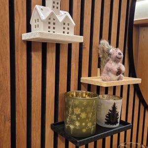 Könnte beinhalten: Präsentation von Dekorationsartikeln auf Regalen vor einer holzgetäfelten Wand. Enthält ein weißes Keramikhaus, ein Plüschhörnchen mit einem Tannenzapfen und zwei Glaskerzenhalter mit Baumdesigns. Die Regale sind cremefarben und schwarz.