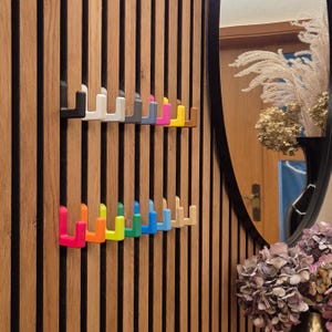Könnte beinhalten: Eine Reihe von U-förmigen Haken in verschiedenen Farben wie Schwarz, Weiß, Grau, Blau, Pink, Gelb, Braun, Orange, Grün und Beige, die an einer vertikalen Wandverkleidung aus Holzlatten mit dunkler Oberfläche zwischen den einzelnen Latten befestigt sind.