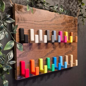 Könnte beinhalten: Ein Wandgarderobe aus Holz mit einer Vielzahl von farbigen Haken. Die Haken sind in zwei Reihen angeordnet und umfassen Farben wie Rot, Orange, Gelb, Grün, Blau und Rosa. Das Holz hat eine natürliche Maserung.