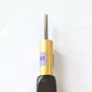 Może przedstawiać: Dłuto ze skóry w kolorze ciemnobrązowym ze złotą metalową tuleją i srebrną igłą. Rękojeść ma mały, okrągły, złoty akcent w pobliżu podstawy. Na tulei widoczna jest fioletowa naklejka z białym tekstem.