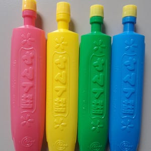 Puede incluir: Cuatro botellas de plástico de colores, rosa, amarillo, verde y azul, cada una con una tapa amarilla. Las botellas tienen texto japonés y diseños florales. Las botellas están dispuestas verticalmente.
