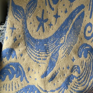 May include: A crocheted blanket with a whale design in light blue on a beige background. The blanket also features stars and waves. The edges have a fringe detail.