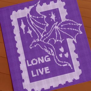 May include: A purple, square, crocheted blanket with a white dragon design inside a postage stamp border. The words "LONG LIVE" are in white block letters below the dragon illustration. The blanket is on a wooden surface.