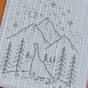 Könnte beinhalten: Kreuzstich-Design mit einer Bergkette, Bäumen, einem Dinosaurier, Mond und Sternen in Grün auf hellblauem Hintergrund. Das Design ist mit einem hellblauen Rand versehen. Der Kreuzstich befindet sich auf einem quadratischen Stoffstück.