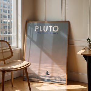 May include: A framed poster with the word "PLUTO" in silver lettering. The poster also includes the text "2.7 BILLION MILES FROM EARTH" and depicts a landscape with a small figure. A wooden chair and table are in the foreground.