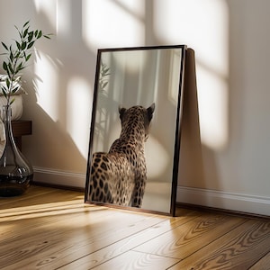 May include: A framed mirror leans against a wall, reflecting a leopard's spotted back and head. The mirror has a dark brown frame. The room has a wooden floor and a vase with a plant.