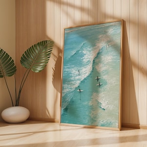 May include: A framed art print featuring a vibrant aerial view of surfers in the ocean. The artwork showcases turquoise waves and several surfers riding the waves. The frame is a light brown color, and the scene is set in a room with a plant.