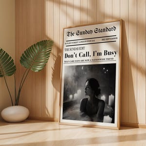 Peut inclure: Une affiche encadrée avec le titre "Don't Call, I'm Busy" dans une mise en page de style journal. L'image présente une photographie en noir et blanc d'une personne dans une baignoire entourée de bougies. L'affiche est affichée contre un mur en bois.