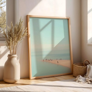 May include: A framed art print featuring a serene beach scene with figures. The artwork is displayed in a light wood frame, leaning against a wall. A beige vase with dried plants and a woven basket with a blanket are nearby.