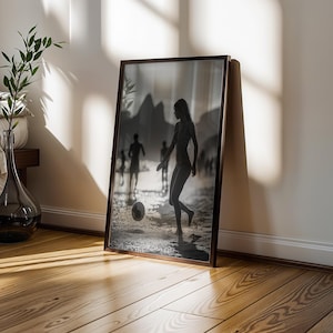 May include: A framed black and white photograph of people playing soccer on a beach. The photograph is leaning against a white wall and a wooden floor. A glass vase with a plant is next to a bed.