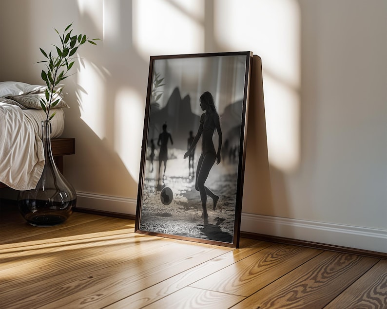 May include: A framed black and white photograph of people playing soccer on a beach. The photograph is leaning against a white wall and a wooden floor. A glass vase with a plant is next to a bed.