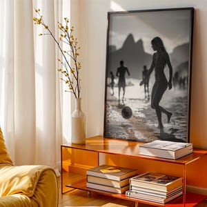 May include: A framed black and white photograph of people playing soccer on a beach is displayed above a modern orange console. A white vase with yellow-flowered branches sits to the left. Books are stacked on the console's shelves, and a yellow armchair is in the foreground.