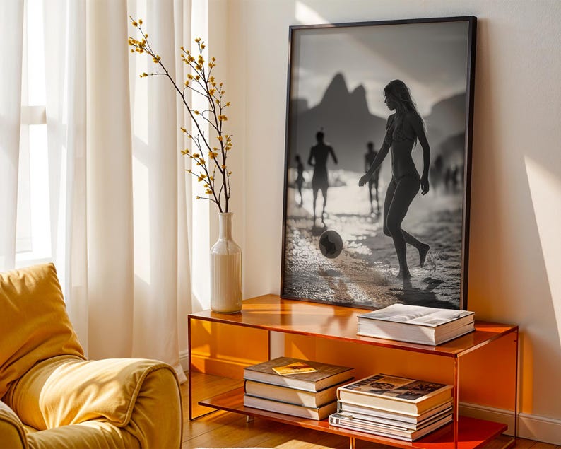 May include: A framed black and white photograph of people playing soccer on a beach is displayed above a modern orange console. A white vase with yellow-flowered branches sits to the left. Books are stacked on the console's shelves, and a yellow armchair is in the foreground.