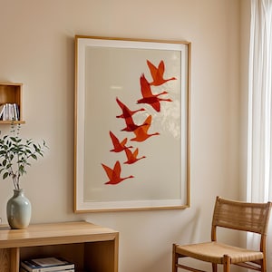 May include: A framed art print featuring a flock of orange birds in flight against a cream background. The artwork is displayed in a room with a wooden table, a chair, and a vase with greenery.