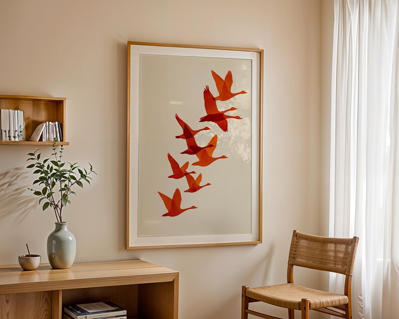 May include: A framed art print featuring a flock of orange birds in flight against a cream background. The artwork is displayed in a room with a wooden table, a chair, and a vase with greenery.