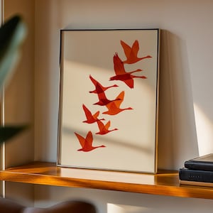 May include: Framed art print featuring a flock of orange geese in flight against a cream background. The artwork is displayed in a gold-toned frame and rests on a wooden shelf. The print adds a touch of nature-inspired decor.