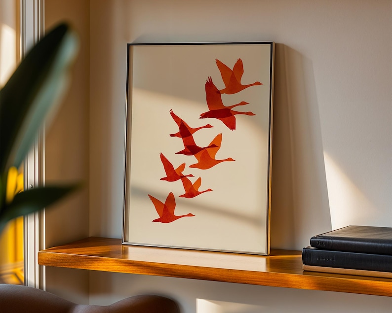 May include: Framed art print featuring a flock of orange geese in flight against a cream background. The artwork is displayed in a gold-toned frame and rests on a wooden shelf. The print adds a touch of nature-inspired decor.