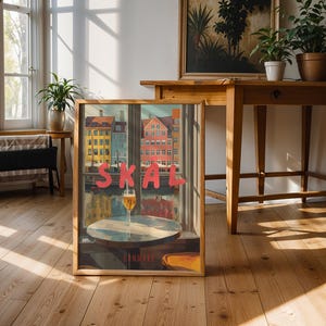 May include: A framed art print featuring a colorful cityscape with the word "SKAL" in red letters. A glass of beer sits on a small table in front of the city view. The word "DANMARK" is at the bottom.