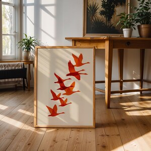 May include: A framed art print featuring a flock of orange birds in flight against a cream background. The artwork is displayed on a wooden floor, leaning against a wall. The print has a natural wood frame.