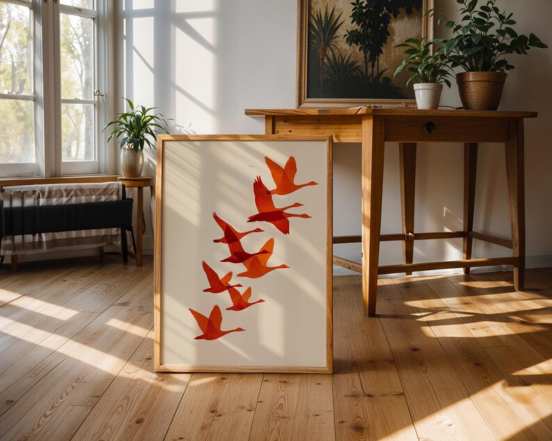 May include: A framed art print featuring a flock of orange birds in flight against a cream background. The artwork is displayed on a wooden floor, leaning against a wall. The print has a natural wood frame.