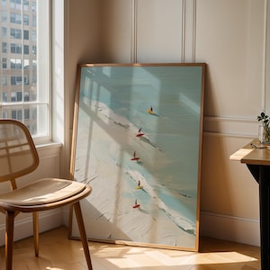 May include: A framed art print depicts a beach scene with surfers. The artwork features a light blue ocean, white waves, and a sandy shore. Five surfers with colorful boards are visible. The frame is a light brown color. A wooden chair and table are in the foreground.
