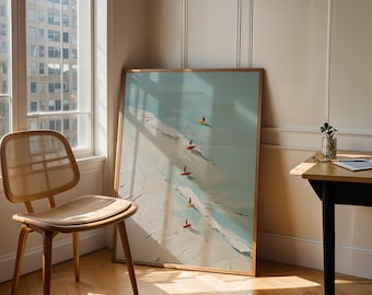 May include: A framed art print depicts a beach scene with surfers. The artwork features a light blue ocean, white waves, and a sandy shore. Five surfers with colorful boards are visible. The frame is a light brown color. A wooden chair and table are in the foreground.