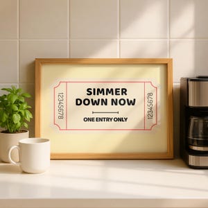 May include: A framed print with the words "SIMMER DOWN NOW" in black text on a ticket-style design. The print is in a light wood frame and sits on a white surface. A coffee maker and a mug are also visible.