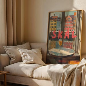 May include: A framed art print featuring a colorful cityscape with the word "SKAL" in large pink letters. A glass of beer sits on a table in front of a window. The print is displayed on a beige sofa with pillows.