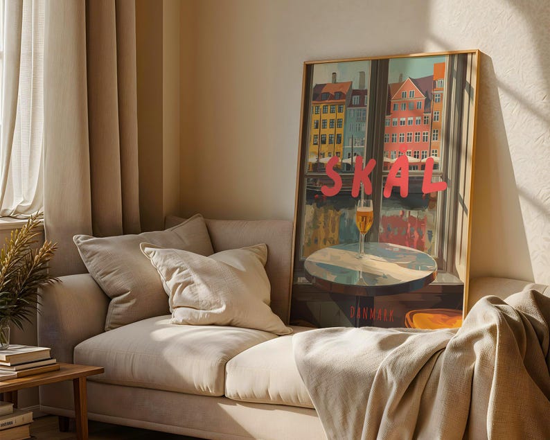 May include: A framed art print featuring a colorful cityscape with the word "SKAL" in large pink letters. A glass of beer sits on a table in front of a window. The print is displayed on a beige sofa with pillows.