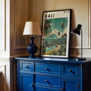 May include: A navy blue dresser with brass knobs supports a framed poster with the word "BALI" and a landscape scene. A navy blue lamp with a beige shade and a black desk lamp are on the dresser.