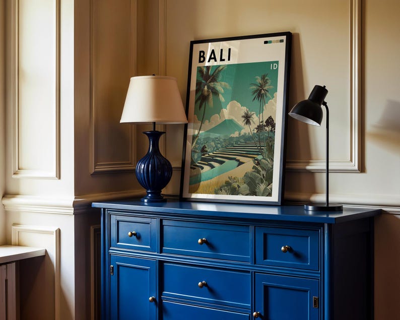 May include: A navy blue dresser with brass knobs supports a framed poster with the word "BALI" and a landscape scene. A navy blue lamp with a beige shade and a black desk lamp are on the dresser.
